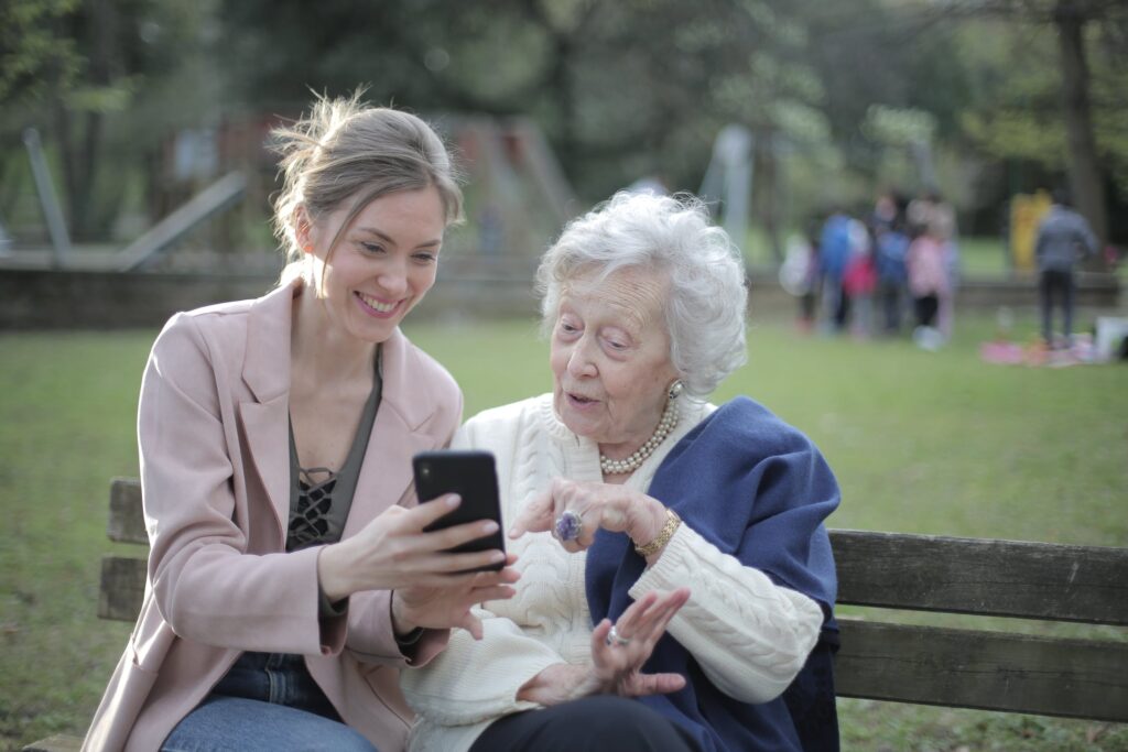 A young women sititng on a bench with an elderly woman, showing her something on a smartphone