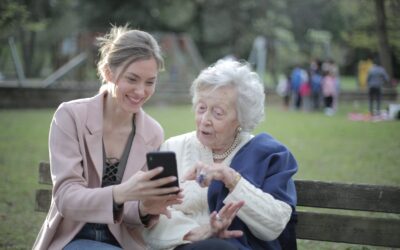 A young women sititng on a bench with an elderly woman, showing her something on a smartphone